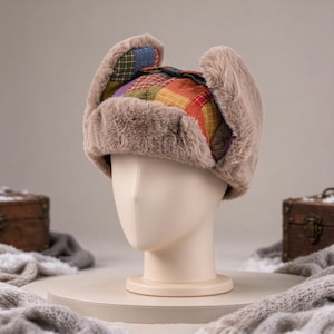 May include: A trapper hat featuring a multi-colored patchwork pattern in shades of red, yellow, green, and blue. The hat is lined with thick, light brown faux fur along the brim and ear flaps, and includes a black plastic buckle strap on top.