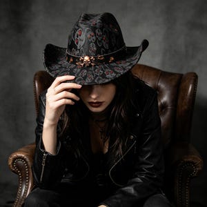 Può includere: Un cappello da cowboy nero con un motivo ripetuto di teschi grigi con occhi rossi. Il cappello include una fascia nera decorata con accenti metallici color rame a forma di teschio e ossa incrociate attorno alla corona e una tesa larga.