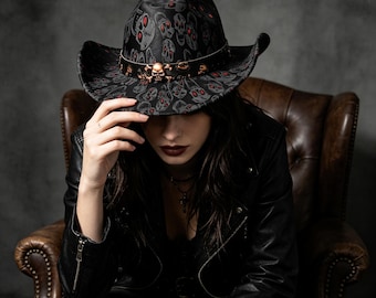 Può includere: Un cappello da cowboy nero con un motivo ripetuto di teschi grigi con occhi rossi. Il cappello include una fascia nera decorata con accenti metallici color rame a forma di teschio e ossa incrociate attorno alla corona e una tesa larga.