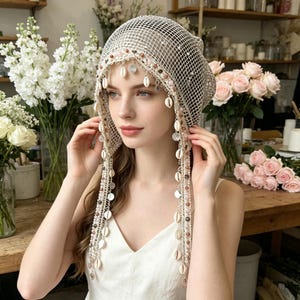 May include: A beige crochet head covering featuring a fine mesh pattern adorned with dangling white cowrie shells, small metallic discs, and multi-colored beads along the front edge and side fringes, worn over a white sleeveless top.