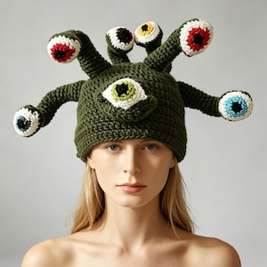 May include: A dark olive green crochet beanie featuring a large central eye and multiple protruding stalks, each topped with a colourful crocheted eye. The eyes have white sclera and pupils in various colours including red, blue, yellow, and green.