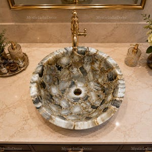 May include: A round vessel sink crafted from polished gray and white agate stone slices with gold-toned veining. The sink features a central drain hole and is paired with a tall, polished gold-toned metal faucet mounted on a beige marble countertop.