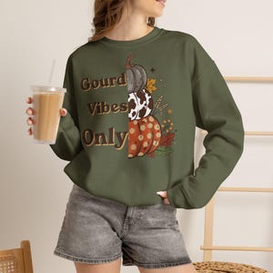Könnte beinhalten: Ein olivgrünes Sweatshirt mit Rundhalsausschnitt und dem Schriftzug Gourd Vibes Only in braunen Buchstaben. Das Design zeigt einen vertikalen Stapel aus drei Kürbissen mit verschiedenen Mustern, darunter Kuhflecken und Punkte, sowie Herbstblätter.
