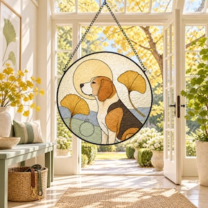 Op de afbeelding: Een ronde glas-in-lood zonnevanger met een beagle in profiel, omringd door gouden ginkgobladeren en een cirkelvormig zonmotief. Het object is gevat in zwart metaal en hangt aan een zwarte ketting voor een helder verlicht raam.
