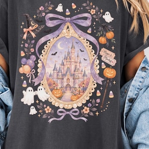 Op de afbeelding: Een antracietgrijs t-shirt met een centraal ovaal kader met een kasteel, pompoenen en vleermuizen. Het ontwerp bevat een paarse strik, spoken, snoepjes en een bordje met de tekst Magic & Mischief, omringd door bloemmotieven.