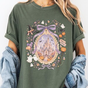 Op de afbeelding: Een donker olijfgroen t-shirt met een centrale afbeelding van een kasteel in een sierlijk ovaal frame. Het ontwerp bevat een paarse strik, spoken, pompoenen, een heksenhoed en een bordje met de tekst Magic & Mischief en een hartje.