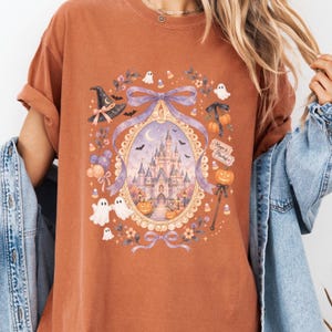 Op de afbeelding: Een roestkleurig t-shirt met een centraal ovaal kader met een kasteelillustratie, omringd door geesten, pompoenen, een heksenhoed en een paarse strik. De tekst "Magic & Mischief" staat op een klein vaantje bij een toverstaf met een pompoen.