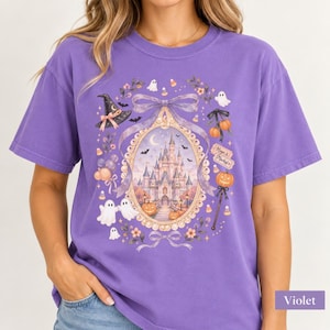 Op de afbeelding: Een violetkleurig T-shirt met korte mouwen en een grafische print van een kasteel in een sierlijk ovaal frame. Het ontwerp bevat spoken, pompoenen, een heksenhoed, strikken en de tekst Magic & Mischief in een feestelijk Halloween-thema.