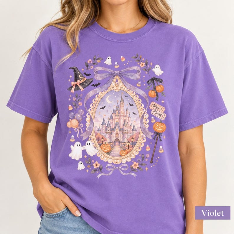 Op de afbeelding: Een violetkleurig T-shirt met korte mouwen en een grafische print van een kasteel in een sierlijk ovaal frame. Het ontwerp bevat spoken, pompoenen, een heksenhoed, strikken en de tekst Magic & Mischief in een feestelijk Halloween-thema.