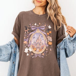 Op de afbeelding: Een donkerbruin t-shirt met korte mouwen en een grafische print van een kasteel in een sierlijke gouden lijst, omringd door geesten, pompoenen, een heksenhoed en een paarse strik. De tekst Magic & Mischief staat op een klein rechthoekig bordje.