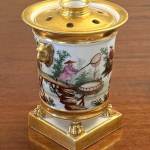 May include: A white porcelain potpourri jar with a gold-trimmed lid featuring multiple circular holes. The cylindrical body is decorated with hand-painted figures in colourful clothing, gold lion head accents, and a square base with gold claw feet.