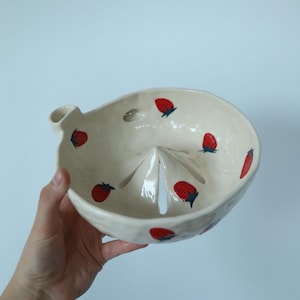 May include: A cream-coloured ceramic berry bowl with a glossy finish, featuring hand-painted red strawberries with green leaves scattered across the interior. The bowl has a built-in drainage spout and a central raised cone for rinsing fruit.