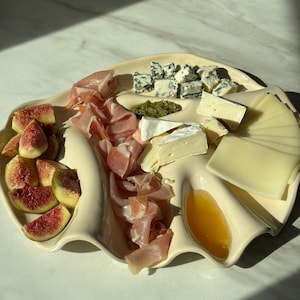 May include: A cream-colored ceramic serving platter with multiple curved compartments holding sliced figs, folded prosciutto, blue cheese crumbles, green pesto, wedges of brie, slices of yellow cheese, and a pool of golden honey.