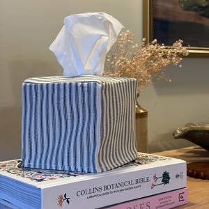 May include: A square fabric tissue box cover featuring a black and white ticking stripe pattern. The cover is constructed with a boxy shape and a top opening, with a single white facial tissue pulled through the centre of the top panel.