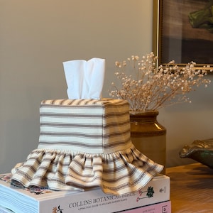 May include: A square fabric tissue box cover featuring a beige and brown horizontal striped pattern with a gathered ruffle trim at the base. The cover is placed over a stack of white facial tissues, resting on top of two stacked hardcover books.
