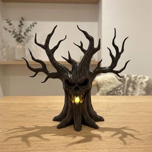 May include: A dark brown, sculpted tree figurine with a menacing, skull-like face and gnarled, spreading branches. The tree features a hollow mouth area containing a small, glowing yellow LED light that illuminates from within the center.