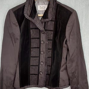 May include: A dark charcoal grey jacket featuring black velvet panels, a ruffled collar, and a front button closure. The interior label reads RoFa MODEN. The jacket has long sleeves and a textured fabric finish, hanging on a wooden hanger.