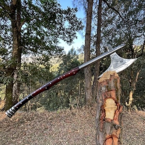 May include: A long-handled battle axe with a polished silver-colored metal head and a dark reddish-brown wooden shaft. The shaft features multiple silver-colored metal rivets and a ribbed silver-colored metal pommel at the base of the handle.