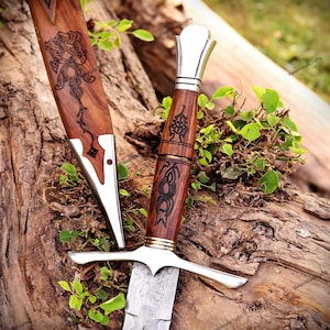 May include: A dagger with a patterned steel blade, a wooden handle featuring dark etched knotwork designs, and a polished silver-coloured pommel and crossguard. The matching wooden scabbard displays similar intricate dark etched knotwork patterns.