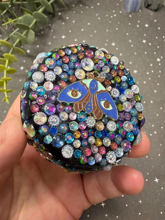 A one-of-a-kind hand-bedazzled moth pill tin — rhinestone-encrusted, witchy, and small enough to hide in a pocket while looking like a tiny piece of art.