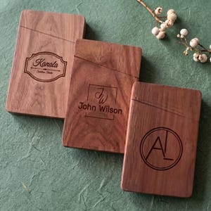 Puede incluir: Tres tarjeteros rectangulares de madera con acabado de veta natural. Cada uno cuenta con una ranura diagonal y diseños grabados con láser: el nombre Kanala, el nombre John Wilson y el monograma AL dentro de un círculo.