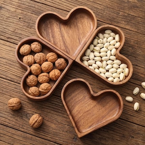 May include: Three heart-shaped wooden bowls with visible grain patterns. One bowl is empty, one contains brown walnuts, and one contains light-colored pistachios. Two additional walnuts are placed outside the bowls on the surface.