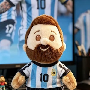 May include: A plush doll featuring a brown beard and hair, wearing a light blue and white vertical striped soccer jersey with the number 10 printed in black on the front and a small yellow and blue emblem on the chest area above the number.