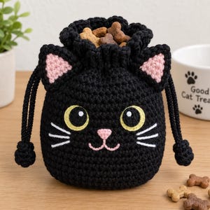 May include: A black crochet drawstring pouch shaped like a cat head with pink inner ears, yellow eyes, white whiskers, and a pink nose. The pouch is filled with brown bone-shaped pet treats and features two black braided cords with knotted ends.
