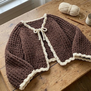 Può includere: Un cardigan all'uncinetto marrone scuro con un bordo smerlato color crema lungo i profili e una chiusura a fiocco coordinata sullo scollo. Il capo presenta maniche lunghe e una lavorazione a maglia grossa e testurizzata.