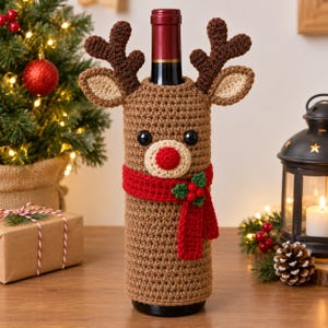 May include: A brown crocheted bottle cover shaped like a reindeer with dark brown antlers, light brown ears, black button eyes, and a cream-colored muzzle with a red nose. A red crocheted scarf with green holly leaves and red berries is attached.
