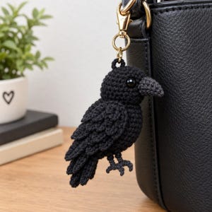 May include: A black crochet raven keychain featuring a textured yarn body, layered wing details, a small dark beak, and a glossy black bead eye. The bird is attached to a gold-toned metal lobster clasp, which is clipped onto a black leather bag.
