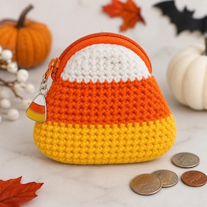 May include: A triangular crochet coin purse designed to resemble a piece of candy corn, featuring horizontal stripes of white, orange, and yellow yarn. The pouch has an orange zipper closure and a small metal charm shaped like candy corn.