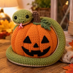 May include: A crocheted orange pumpkin with a black felt jack-o'-lantern face, featuring a brown stem and a green leaf, wrapped in a long, coiled green crocheted snake with black eyes and a small pink tongue protruding from its mouth.