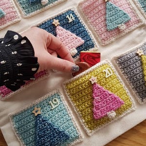 May include: A fabric advent calendar featuring square crochet pockets in various colors including blue, pink, and yellow. Each pocket displays a crocheted Christmas tree, a wooden star, and a number. A hand is placing a small chocolate into pocket 14.