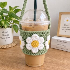 Puede incluir: Un vaso de plástico transparente para café helado con pajita negra, envuelto en una funda de ganchillo verde salvia con aplicaciones de margaritas blancas tridimensionales con centros amarillos y una correa de ganchillo a juego.