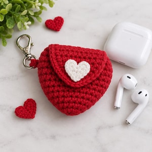 May include: A red heart-shaped crochet case for wireless earbuds featuring a white crocheted heart appliqué on the front flap and a silver metal lobster clasp keychain. The case is accompanied by a white wireless charging case and two earbuds.