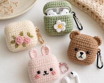 Op de afbeelding: Vier gehaakte AirPod-hoesjes: een groene hoes met een witte bloem, een crèmekleurige met roze rozen, een lichtroze konijnenhoes met oren en een bruine berenhoes met oren. Elk hoesje is voorzien van een metalen karabijnhaak of ring.