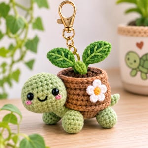 Puede incluir: Un llavero de tortuga de ganchillo que presenta una tortuga de color verde claro con ojos negros y mejillas rosadas. La tortuga lleva una maceta de ganchillo marrón en su espalda con una planta verde. Una flor blanca con centro amarillo está en la maceta.