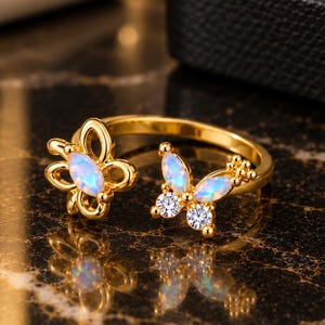 May include: A gold-toned open ring featuring two butterfly-shaped motifs. One butterfly is accented with a marquise-cut iridescent opal-like stone, while the other features a marquise-cut opal-like stone paired with two small round clear stones.