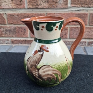 May include: A ceramic jug with a cream-coloured base, featuring a hand-painted brown rooster standing in green grass. The rim and handle are a solid brown, with dark green decorative accents around the neck and a glossy, crackled glaze finish.