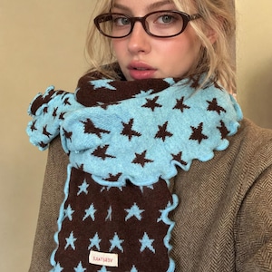 May include: A knit scarf featuring a reversible pattern of brown stars on a light blue background and light blue stars on a brown background. The scarf has a ruffled edge and a small rectangular white label with the text AEBELVITE.