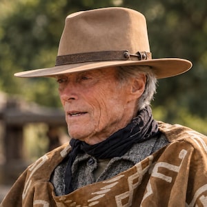 Può includere: Un cappello fedora in feltro color marrone chiaro con una fascia in pelle marrone, indossato con un fazzoletto da collo nero e un poncho marrone con un motivo geometrico bianco. Il capo è sovrapposto a una camicia grigia testurizzata.