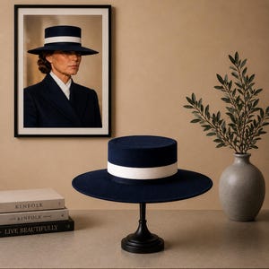 May include: A navy blue flat-top hat with a wide brim and a thick white ribbon band around the crown. The hat is displayed on a black pedestal stand. The design features a structured silhouette with a smooth, felt-like texture and clean lines.