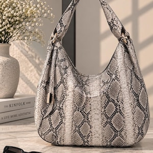 Könnte beinhalten: Eine Hobo-Schultertasche mit grau-weißem Schlangenledermuster. Die Tasche verfügt über einen einzelnen integrierten Griff, der mit goldfarbenen Metallringen befestigt ist, sowie eine dekorative Quaste mit goldfarbener Metallkappe an der Seite.