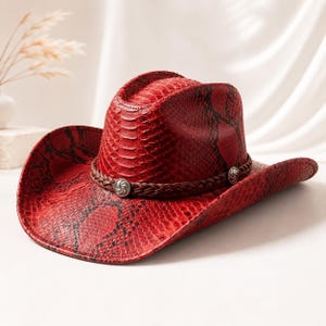 May include: A red cowboy hat featuring a textured snakeskin pattern with black markings. The hat includes a dark brown braided band around the crown, accented with two circular silver-toned metal conchos with a spiral design on the sides.