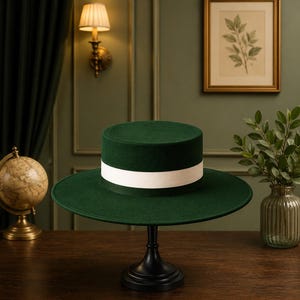 Puede incluir: Un sombrero de color verde oscuro con la parte superior plana, ala ancha circular y una textura lisa y sólida. El sombrero presenta una banda de cinta blanca ancha y contrastante envuelta horizontalmente alrededor de la base.