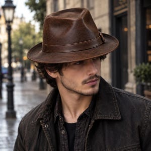 Puede incluir: Un sombrero fedora de color marrón con una textura similar al cuero desgastado y una banda marrón a juego alrededor de la copa. El sombrero presenta una parte superior pellizcada y un ala estructurada, ofreciendo una silueta clásica.