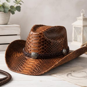 May include: A brown cowboy hat featuring a textured snakeskin pattern across the crown and brim. The hat is accented with a dark brown braided leather band that includes three circular silver-toned metal conchos with intricate floral engravings.