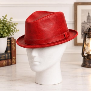 Pode incluir: Um chapéu fedora vermelho vibrante com uma textura lisa ligeiramente desgastada e uma faixa vermelha a condizer à volta da copa. O chapéu é exibido numa cabeça de manequim branca, destacando a sua aba estruturada e design clássico.