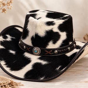 Op de afbeelding: Een cowboyhoed met een zwart-wit koeienvlekkenpatroon en een textuur van echt haar. De hoed is voorzien van een donkerbruine leren band versierd met zilverkleurige metalen studs, gevlochten lederen accenten en een centrale turkoois steen.